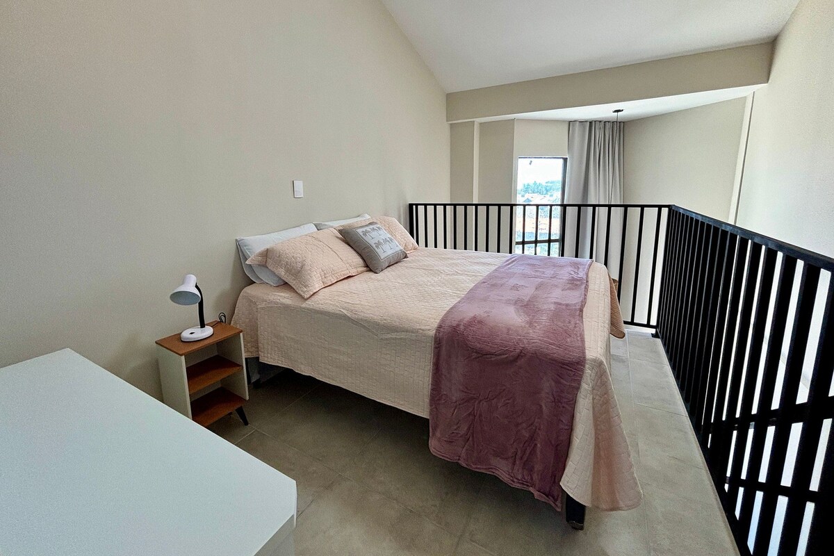 A bedroom with a bed, bedside table, lamp, and a view of the outside.