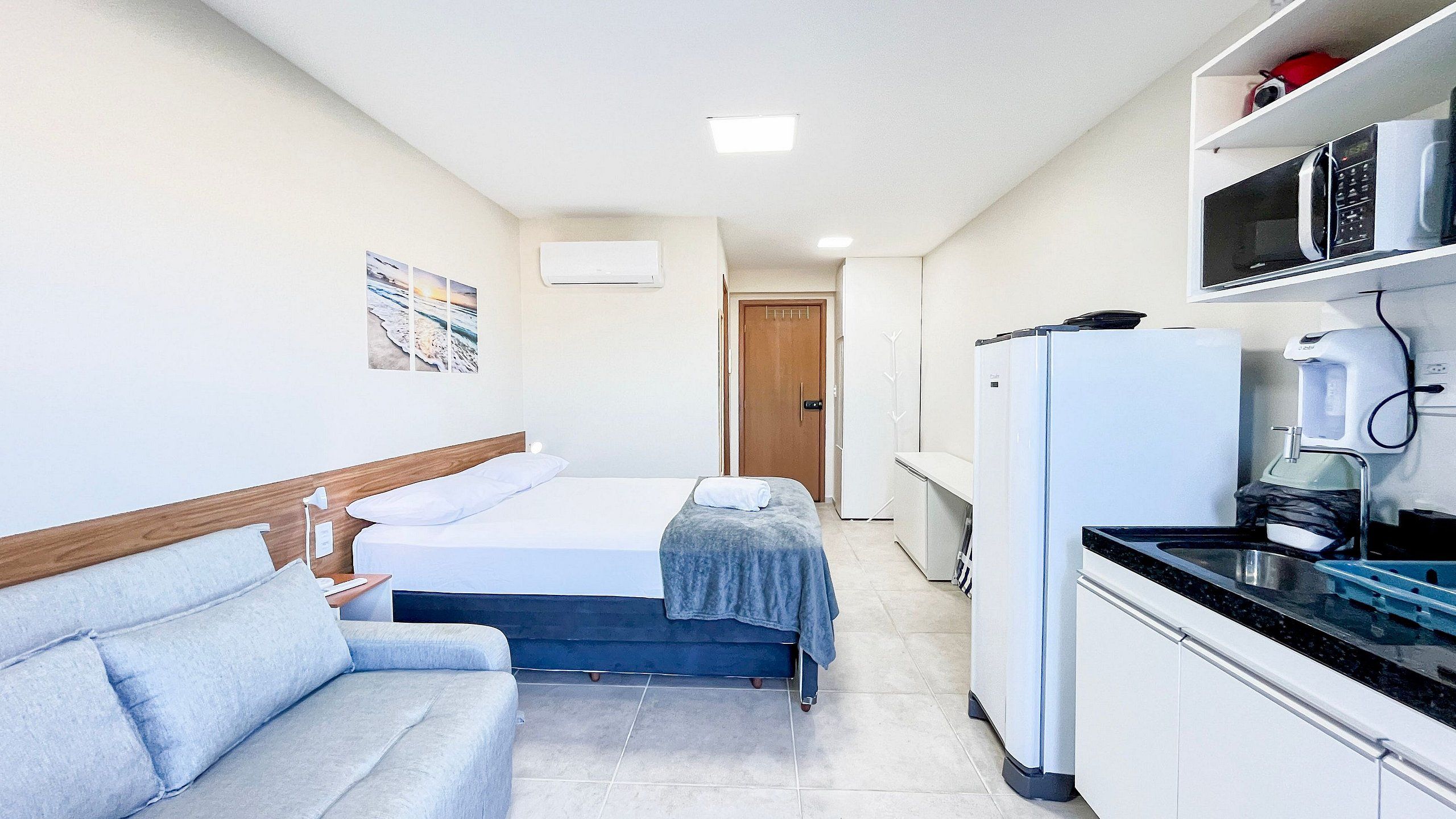 A compact hotel room with a bed, a sofa, a kitchenette, and a door leading to another room.