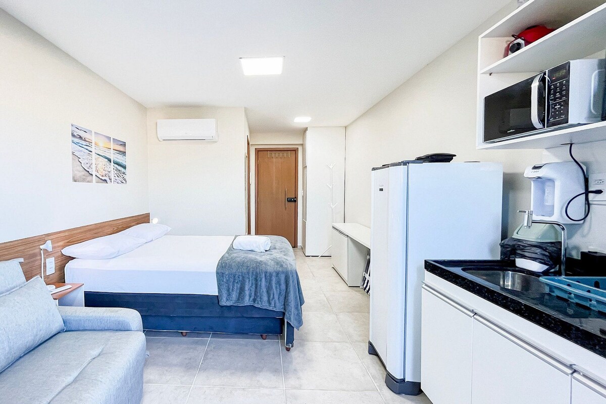 A compact hotel room with a bed, a sofa, a kitchenette, and a door leading to another room.