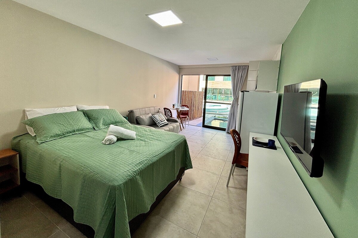 A hotel room with a bed, a TV, a dining area, and a view of the outdoors.