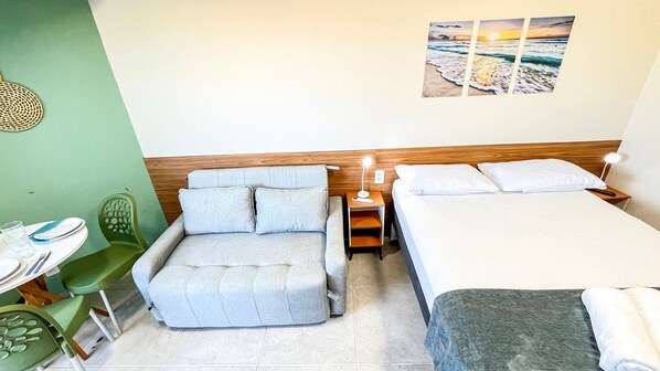 A hotel room with a bed, a sofa, a small table with a chair, and a wall-mounted picture. - Max Carneiros Suites - Com e sem Piscina Privativa (Tamandaré)