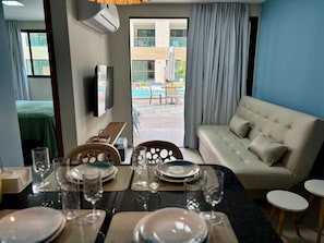 Habitación de hotel con una mesa de comedor para dos personas, un sofá, un televisor y vistas a un área de piscina al aire libre.