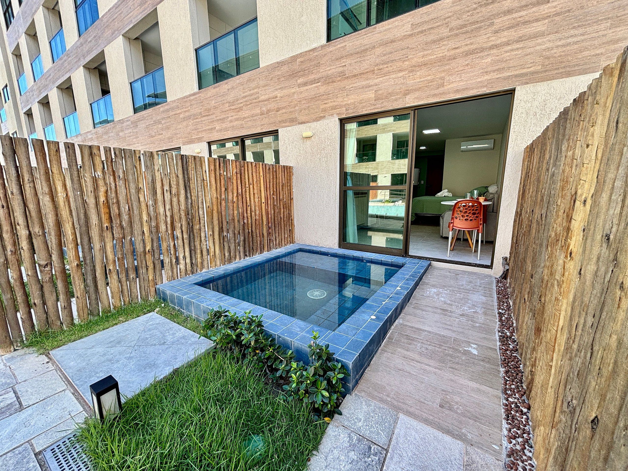 A modern outdoor swimming pool with a wooden fence, a small garden area, and a view of a building with large windows.