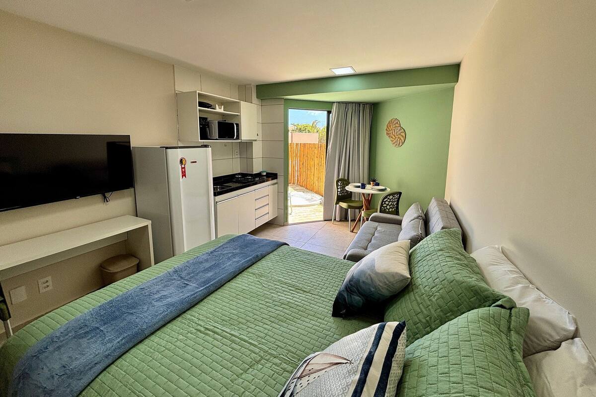 Un dormitorio con cama, televisión, refrigerador, una pequeña mesa con sillas y vistas al exterior.