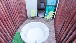 Un jacuzzi en un espacio exterior estrecho con cerco de madera, dos sillas y una vista parcial de un dormitorio con cama y ventana.