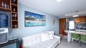 A living room with a white sofa, a painting of a beach scene, a water dispenser, and a kitchen area with a refrigerator and a hanging light fixture. - Max Carneiros Suites - Com e sem Piscina Privativa (Tamandaré)