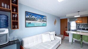 Un salon avec un canapé blanc, un tableau représentant une scène de plage, un distributeur d’eau et un coin cuisine équipé d’un réfrigérateur et d’un luminaire suspendu.