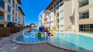 A swimming pool with a slide and a play structure, surrounded by multi-story buildings with balconies. - Max Carneiros Suites - Com e sem Piscina Privativa (Tamandaré)