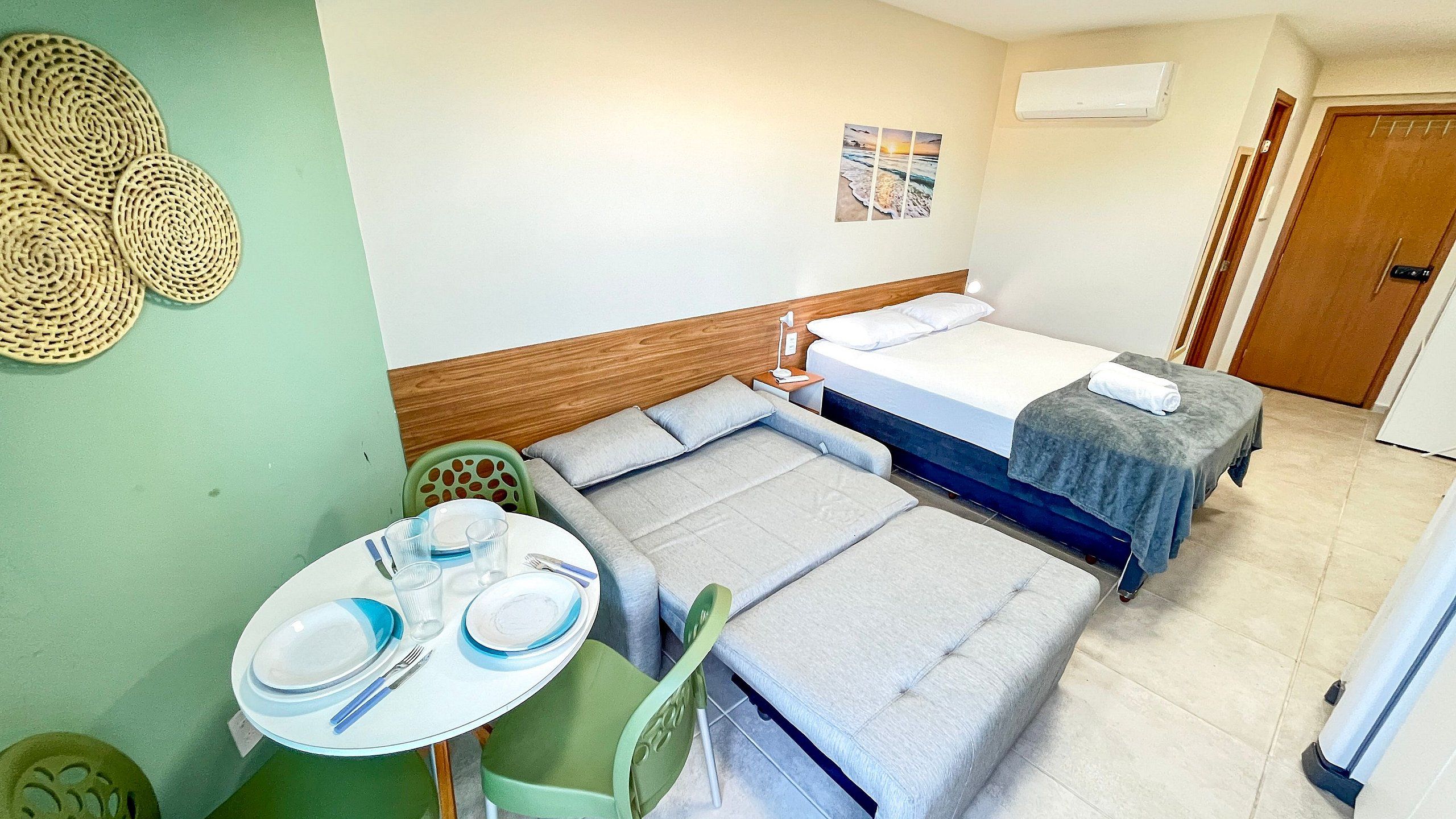 Quarto de hotel com duas camas, uma mesinha com pratos e talheres, uma cadeira verde e um ar condicionado fixado na parede.