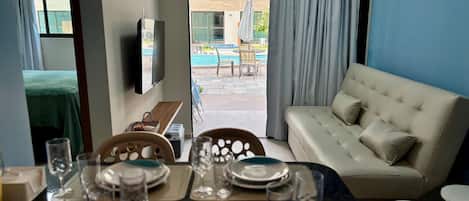 A hotel room with a dining table set for two, a sofa, a TV, and a view of an outdoor pool area.