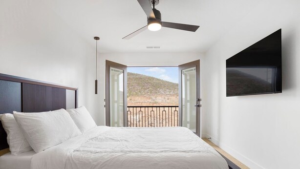 Een slaapkamer met een bed, een plafondventilator, een aan de muur bevestigde televisie en een balkon met uitzicht op een rotsachtig landschap.