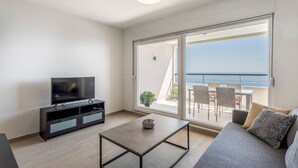 Una sala de estar moderna con un televisor de pantalla plana, una mesa de centro y un sofá. Hay un balcón con vistas al mar.