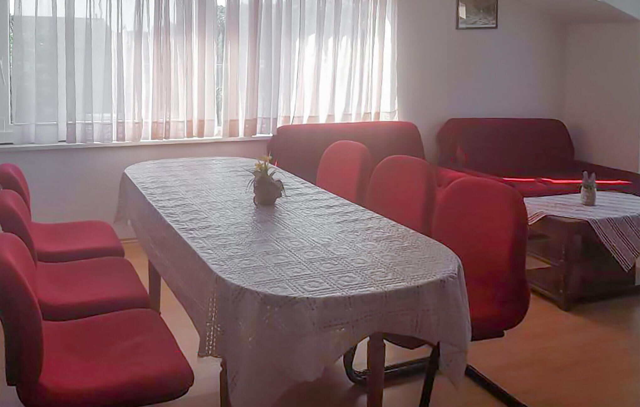 A conference room with a long table, red chairs, a small table with a vase, and a window with sheer curtains.