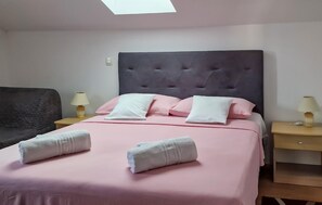 Ein ordentlich bezogenes Bett mit rosa Bettwäsche und weißen Kissen, zwei Nachttische mit Lampen sowie ein dunkel gepolsterter Sessel.