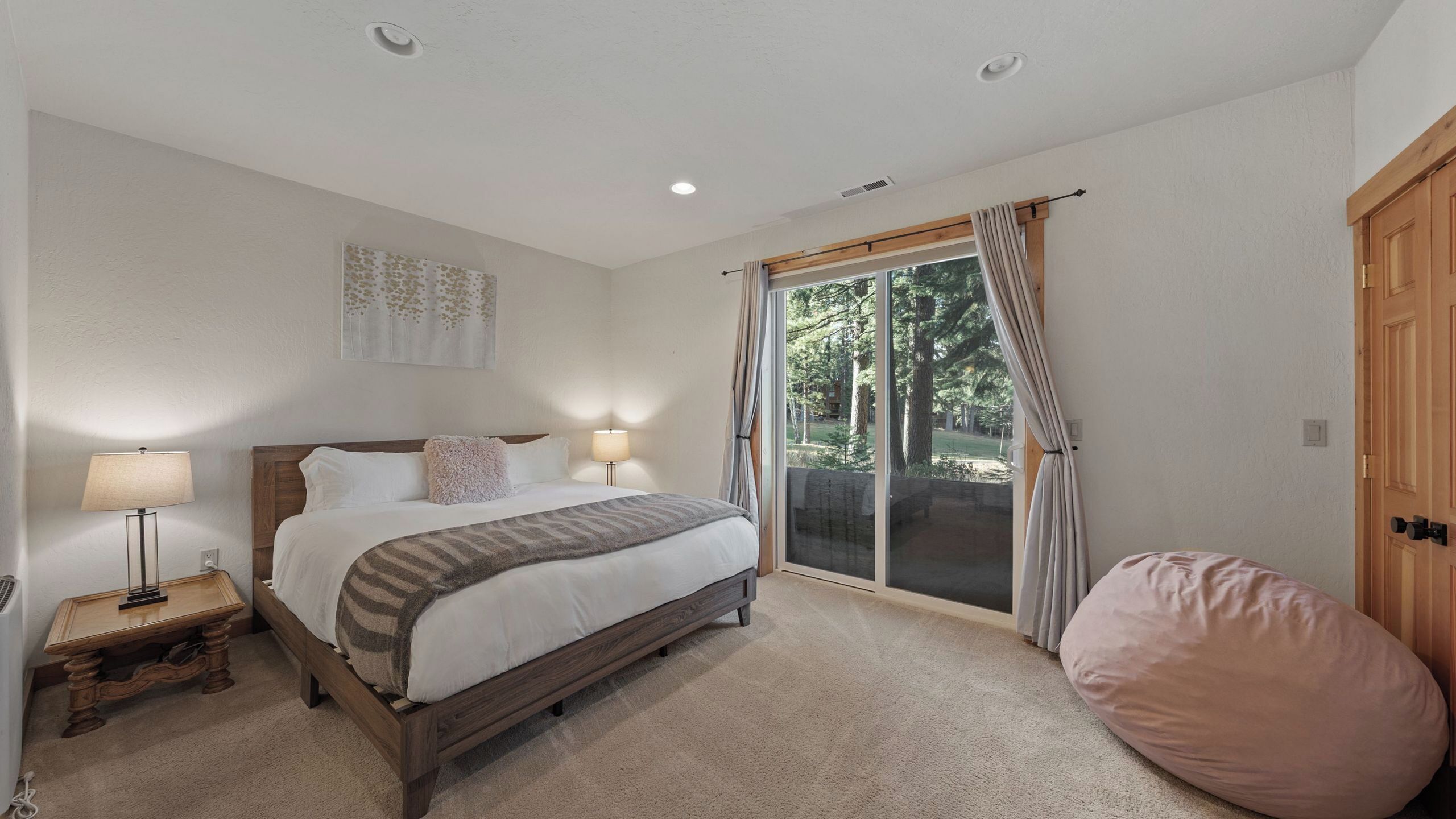 A bedroom with a bed, bedside lamps, a wooden nightstand, a large window with curtains, and a sliding glass door leading to an outdoor area.