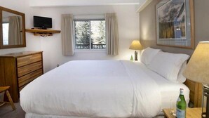 A hotel room with a bed, a television on a wooden shelf, a window with curtains, a painting on the wall, and a bedside table with a bottle. - Amazing Lionshead 2 bd/2ba condo. Walk to slopes and shops. Amazing Hot Tub! (Vail)