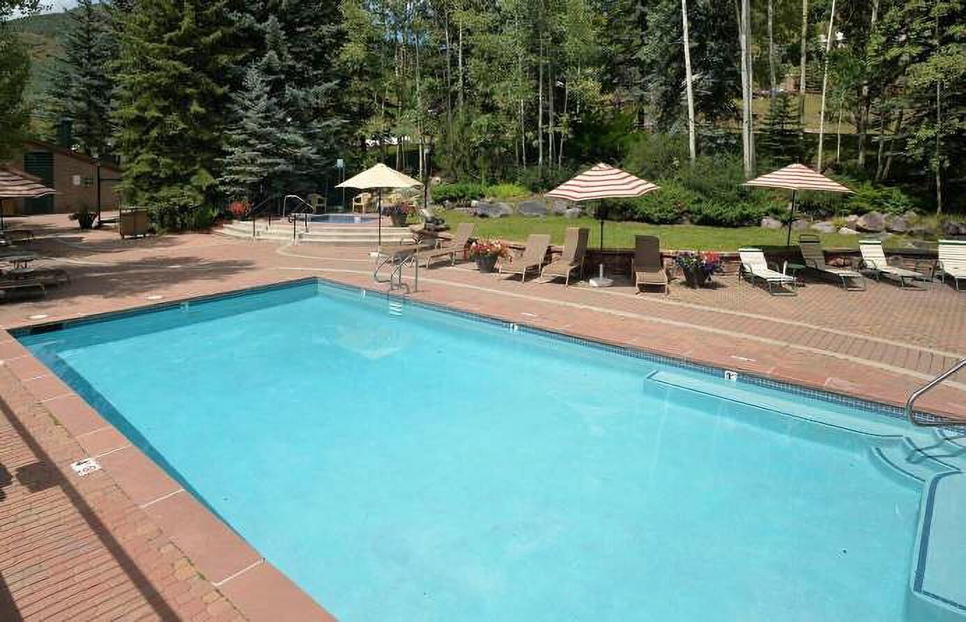 A rectangular swimming pool with a brick border, surrounded by lounge chairs and umbrellas, set in a wooded area.