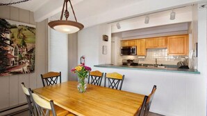 A dining area with a wooden table and chairs, a vase with flowers, and a kitchen with wooden cabinets and a glass countertop. - Amazing Lionshead 2 bd/2ba condo. Walk to slopes and shops. Amazing Hot Tub! (Vail)