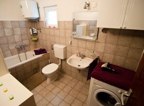 Ein kleines Badezimmer mit Toilette, Waschbecken und Badewanne. Über dem Waschbecken befindet sich ein Fenster und in der Ecke steht eine Waschmaschine.