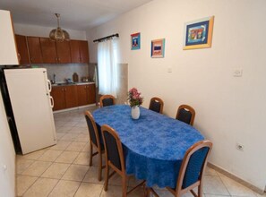 Ein Essbereich mit einem runden Tisch, der mit einer blauen Tischdecke bedeckt ist und von Holzstühlen umgeben wird. Links befindet sich ein weißer Kühlschrank und im Hintergrund hängen Holzausziehtische.