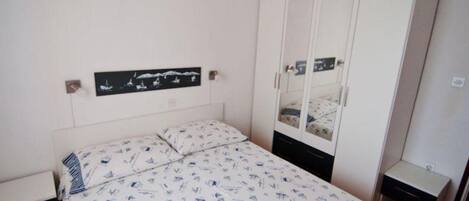 A bedroom with a bed, a wardrobe, a nightstand, and a wall-mounted shelf with a picture.