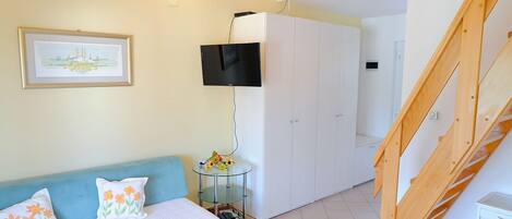 A room with a blue sofa, a white wardrobe, a TV mounted on the wall, and a wooden staircase.