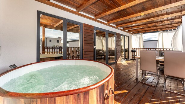 Una terraza de madera con un jacuzzi, mobiliario exterior y vistas a edificios al fondo.