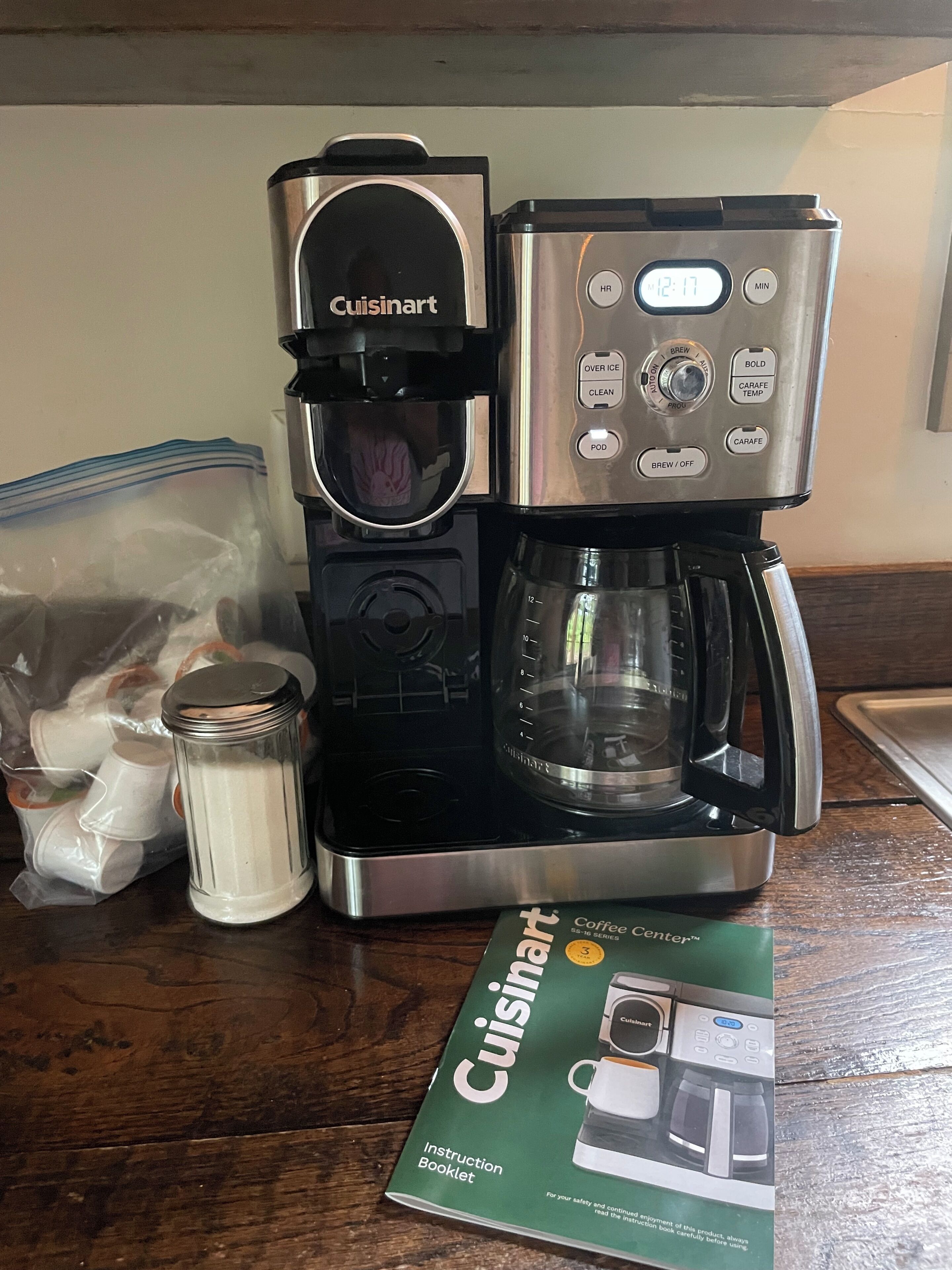 Cuisinart-kahvinkeitin, jossa on kahvikapseliteline, lasinen kahvikannu ja Cuisinart-käyttöohje, kaikki puupöydällä.