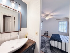 Une salle de bain avec un lavabo blanc, un meuble-lavabo en bois et un miroir avec éclairage intégré. On y voit également une chambre à coucher avec un lit, un ventilateur de plafond et une fenêtre.
