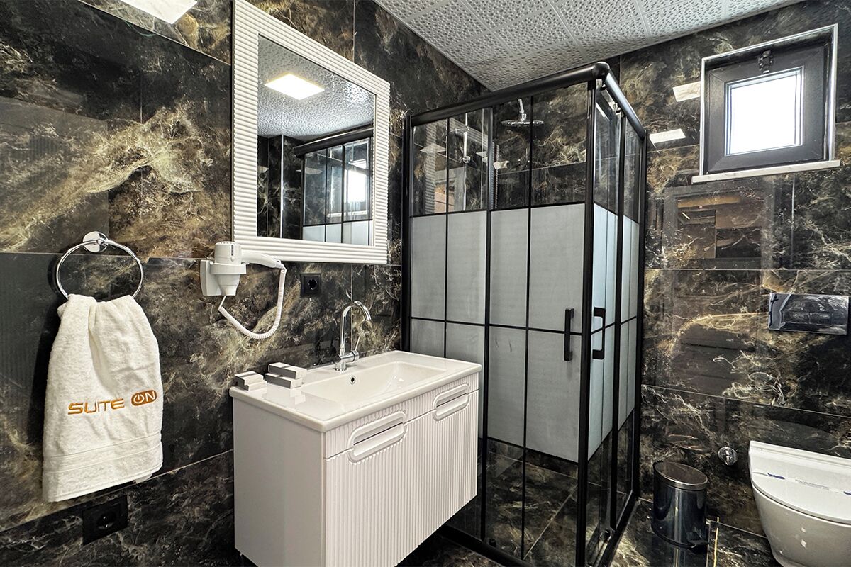 A modern bathroom with a marble wall, a white sink, a glass shower enclosure, and a towel rack with a towel labeled "SUITE ON".