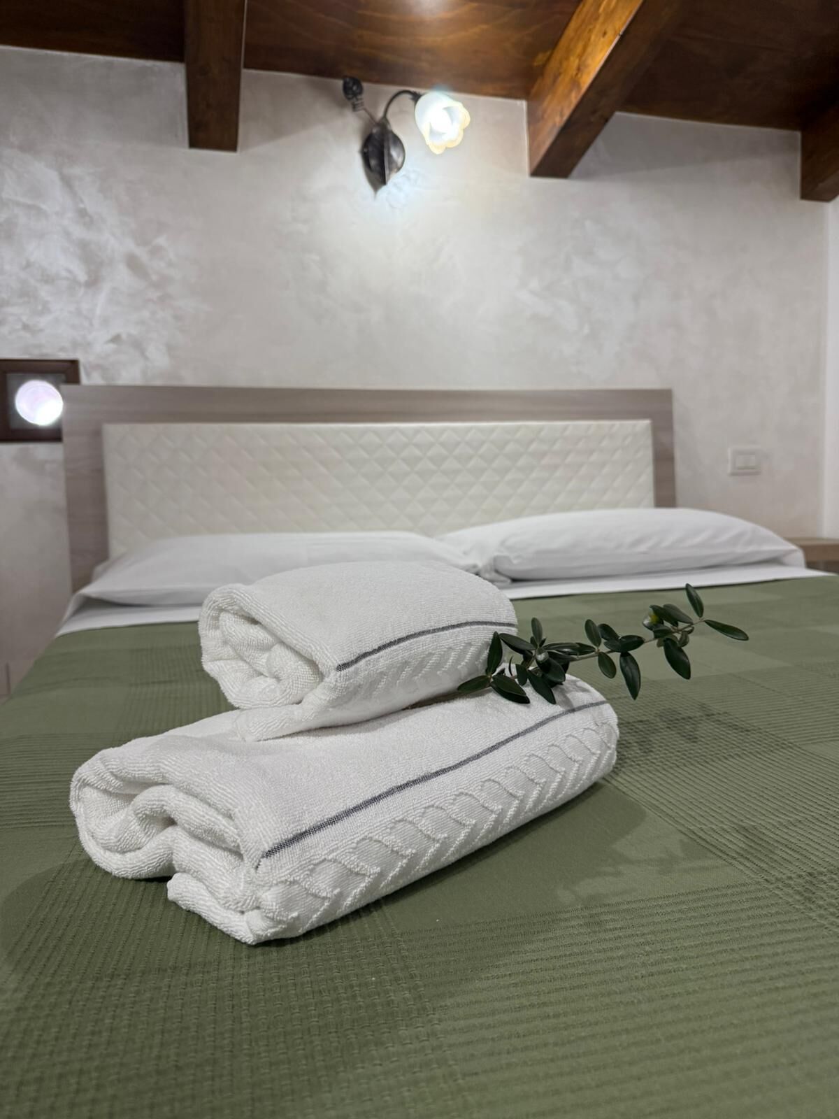 A neatly made bed with white linens and a green bedspread, a decorative wall light, and a plant on the bed.