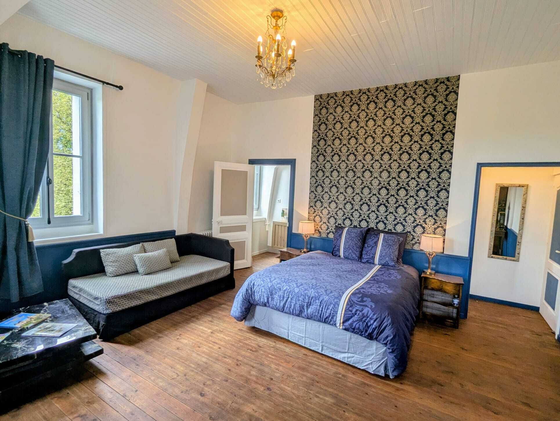 Een slaapkamer met een bed, een bank, een klein tafeltje, een kroonluchter en behang met een patroon.