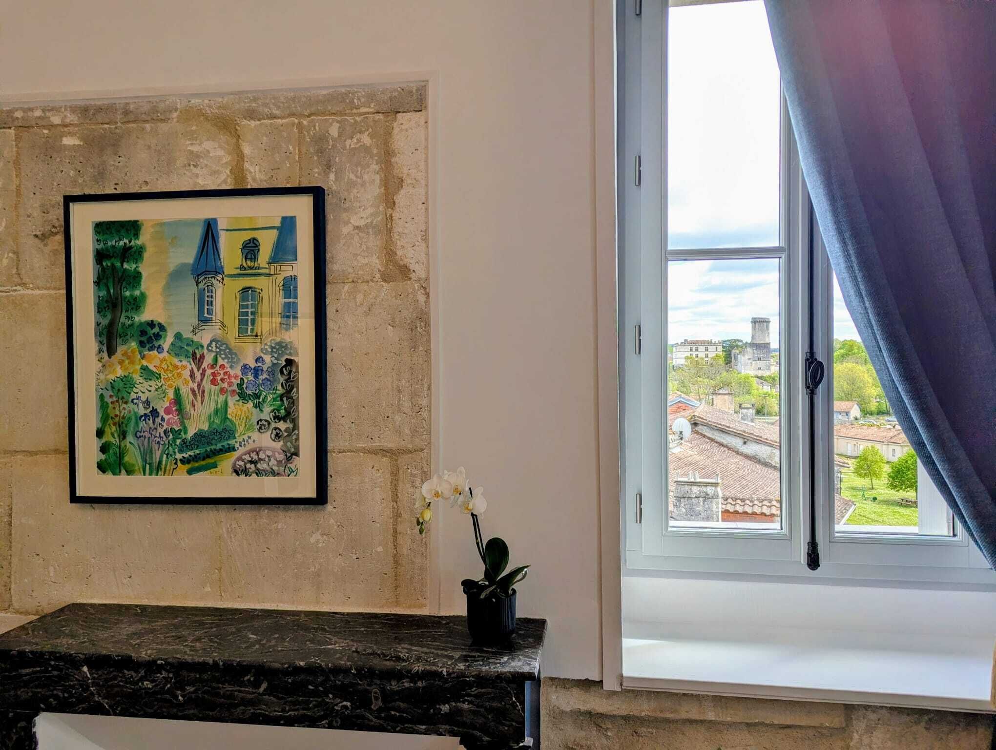 Een kamer met een schilderij in lijst van een kleurrijk landschap, een raam met uitzicht op gebouwen, en een tafeltje met een plant in pot.