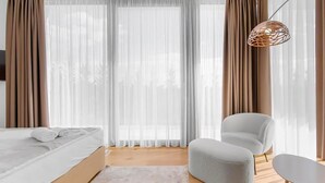 Ein modernes Schlafzimmer mit einem Bett, einem grauen Sessel, einem kleinen Tisch und transparenten Vorhängen.