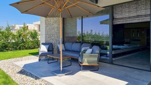 Ein Sitzbereich im Freien mit einer Couch, Stühlen und einem Tisch unter einem großen Sonnenschirm, direkt neben einer Glastür, die zu einem modernen Innenbereich führt.