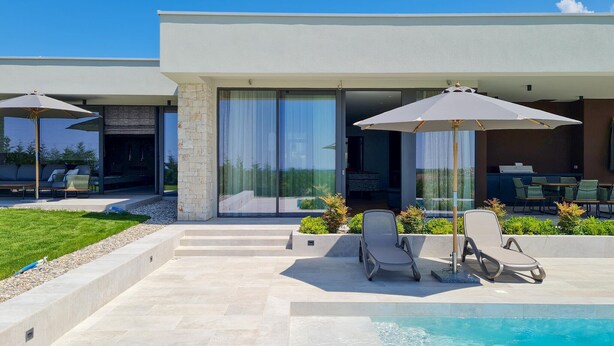 Ein moderner Poolbereich im Freien mit Liegestühlen und Sonnenschirmen, einer Steinsäule und Blick auf den Pool durch große Schiebetüren.