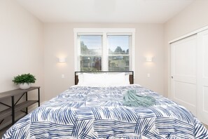 Een slaapkamer met een bed, een raam met zonwering, een nachtkastje met een plant en een deur.
