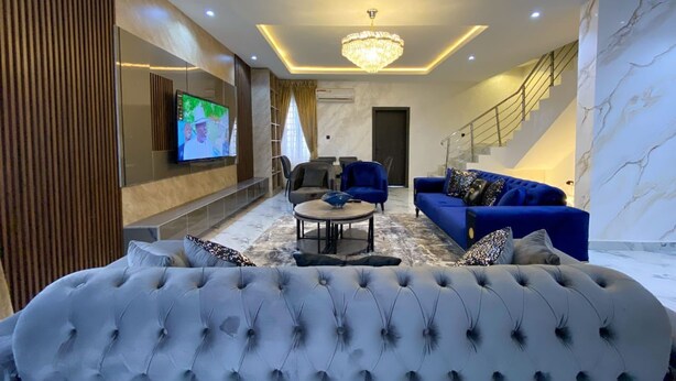 Uma sala de estar moderna com um sofá grande e estofado, um sofá azul, uma mesa de centro redonda e uma televisão montada na parede.