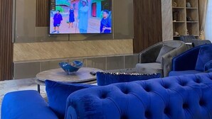 Uma sala de estar moderna com uma grande televisão de tela plana fixada na parede, um sofá azul macio, uma mesa de centro redonda e um lustre decorativo.