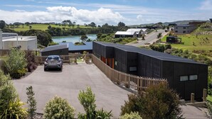 A modern building with a car parked in front, surrounded by greenery and a view of a lake. - Modern Family-Friendly Sunny Raglan Bach with Spa Pool, BBQ & Large Deck (Raglan)
