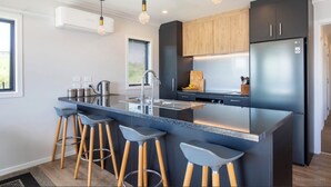 A modern kitchen with a central island, bar stools, stainless steel appliances, and wooden cabinets. - Modern Family-Friendly Sunny Raglan Bach with Spa Pool, BBQ & Large Deck (Raglan)