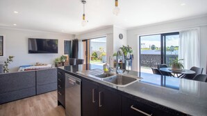 A modern kitchen with a central island, built-in dishwasher, and a large window overlooking a garden. - Modern Family-Friendly Sunny Raglan Bach with Spa Pool, BBQ & Large Deck (Raglan)