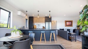 A modern kitchen with a central island, bar stools, and a dining area with a table and chairs. - Modern Family-Friendly Sunny Raglan Bach with Spa Pool, BBQ & Large Deck (Raglan)