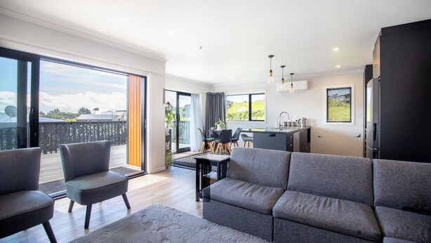 A modern living room with a grey sofa, two armchairs, and a dining area with a table and chairs. - Modern Family-Friendly Sunny Raglan Bach with Spa Pool, BBQ & Large Deck (Raglan)