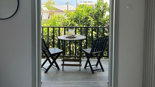 Un balcon avec une table et des chaises, un climatiseur fixé au mur, et une vue sur la verdure et les bâtiments en arrière-plan.
