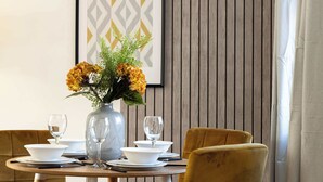 A dining table set with white plates, bowls, and wine glasses, a vase with yellow flowers, and a framed geometric artwork on the wall.
