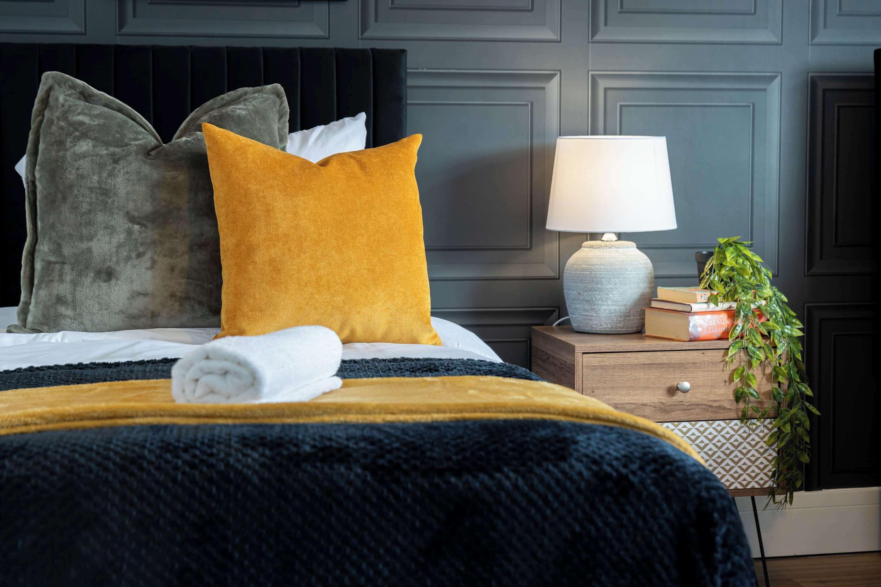 Een net opgemaakt bed met een zwart hoofdeinde, een mosterdkleurig kussen, een grijs kussen en een witte handdoek. Er staat een nachtkastje met een lamp, een boek en een potplantje bij.