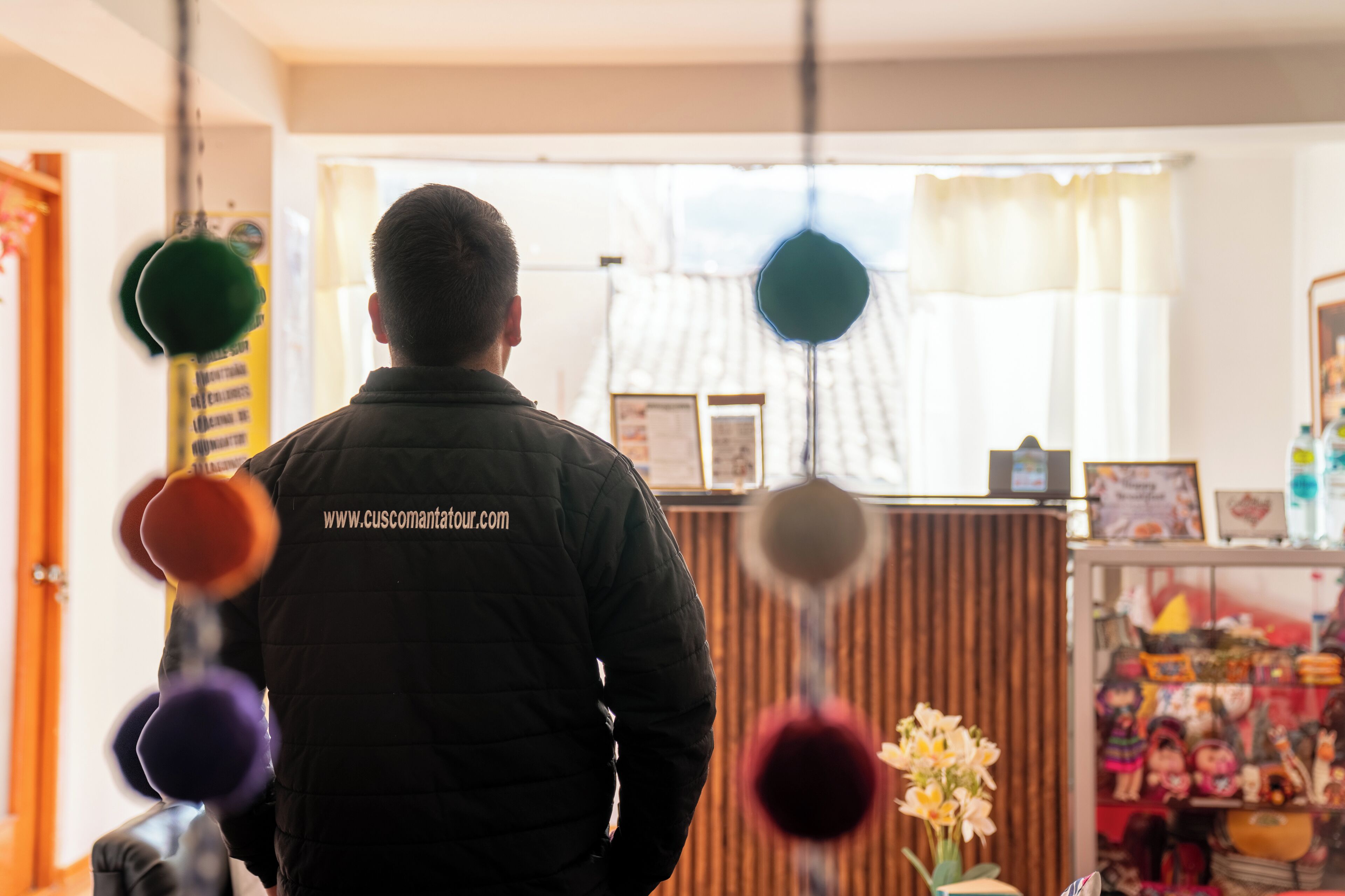 A person in a black jacket with a website URL on the back, standing in a room with a wooden counter and various items on display.