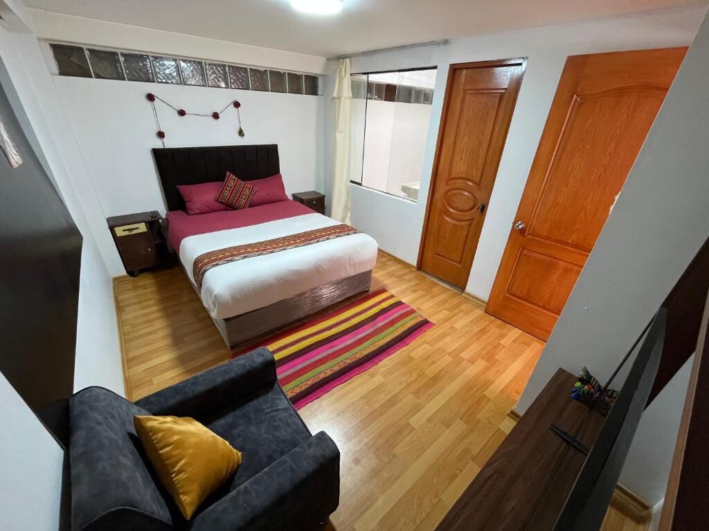 Un dormitorio con cama, mesita de noche, piso de madera, ventana y puerta.