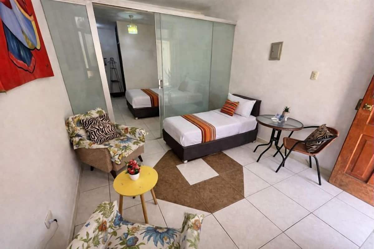 A hotel room with a bed, a sofa, a small table, and a chair. There is a sliding door leading to another room.